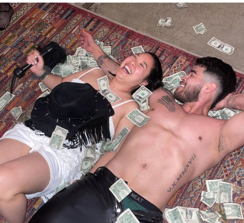 Two people laughing on a colorful patterned rug amid scattered one-dollar bills, a shirtless tattooed man and a woman in white shorts holding a microphone with a black fringed hat nearby — playful indoor party scene.