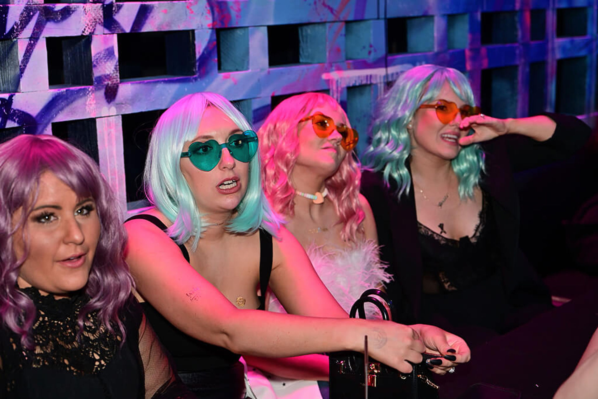 Four people in pastel wigs and heart-shaped sunglasses partying in a neon-lit nightclub booth, wearing stylish party outfits and holding a small handbag