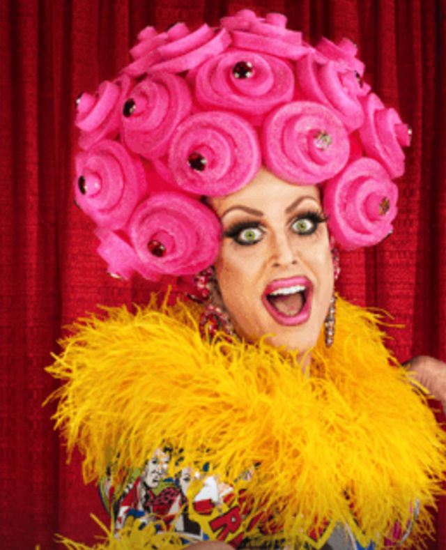 Energetic drag performer with a jewel-studded pink rose wig, dramatic makeup and a yellow feather boa against a red theater curtain.