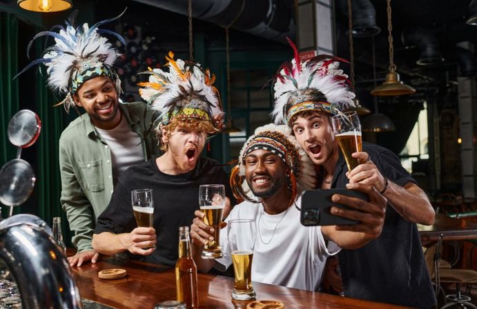 Four friends wearing colorful feathered headdresses cheer and take a selfie while raising beer glasses at a lively bar.