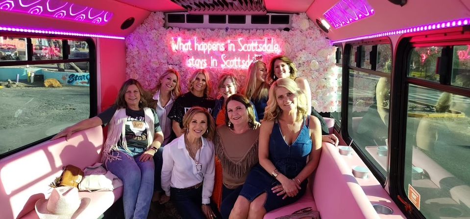 Smiling group of women posing inside a pink party bus with a flower-covered wall and neon sign reading “What happens in Scottsdale stays in Scottsdale,” pink LED lighting and large windows