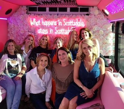 Group of smiling women posing inside a pink party bus with plush benches, floral wall and neon sign reading "What happens in Scottsdale stays in Scottsdale", sunlight streaming through the windows for a celebratory vibe.
