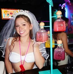 Smiling bride-to-be in a veil and white crop top at a lively bar bachelorette party, holding a pink cocktail from a rotating display of square glass bottles with wooden stoppers.