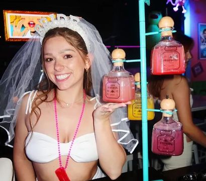 Smiling bride-to-be in a veil and white crop top at a lively bar bachelorette party, holding a pink cocktail from a rotating display of square glass bottles with wooden stoppers.