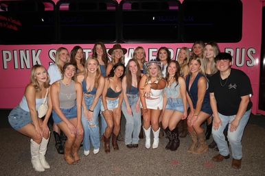 Large group photo at night in front of a bright pink party bus, about 18 friends smiling and posing in denim, shorts and cowboy boots, with one person in a white outfit and cowboy hat front-center — lively girls’ night out vibe