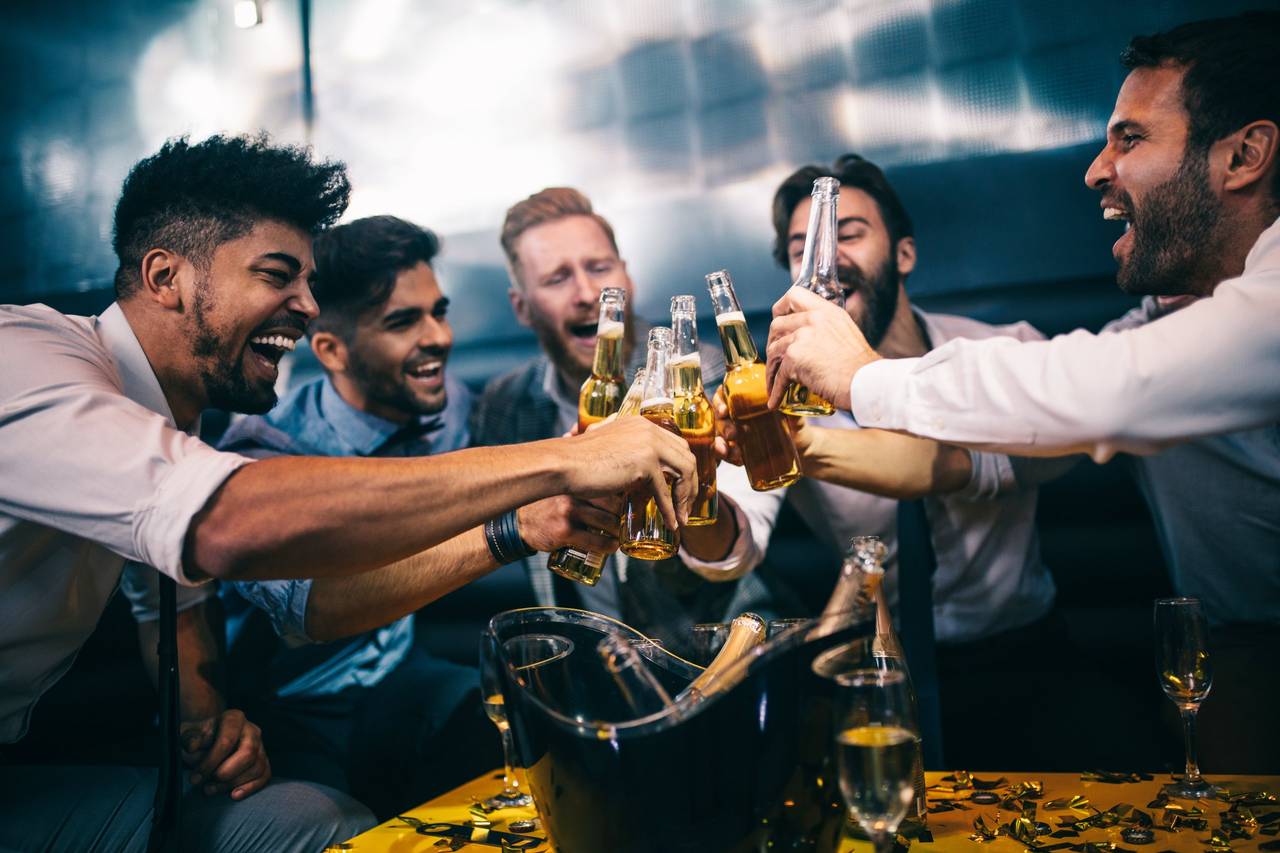 Six friends clinking beer bottles and cheering in a dim modern bar lounge, a celebratory toast around a table with an ice bucket and confetti