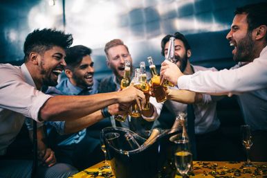Six friends clinking beer bottles and cheering in a dim modern bar lounge, a celebratory toast around a table with an ice bucket and confetti