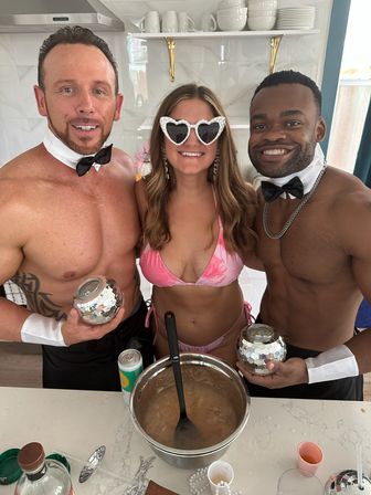 Kitchen party: woman in pink bikini and heart-shaped sunglasses between two shirtless bow-tie servers holding mirrored disco-ball votives over a punch bowl on a countertop with drinks and glassware