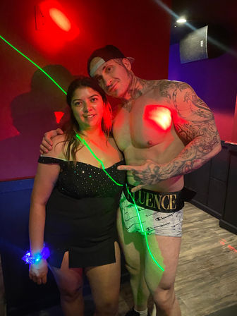 Woman in a black party dress with glowing wristband posing with a shirtless, heavily tattooed male dancer on a club dance floor lit by red and green laser lights.