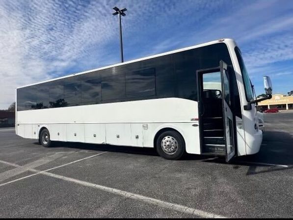 White coach bus with dark tinted windows parked in a suburban shopping-center parking lot, front passenger door open under a bright blue sky with scattered clouds.