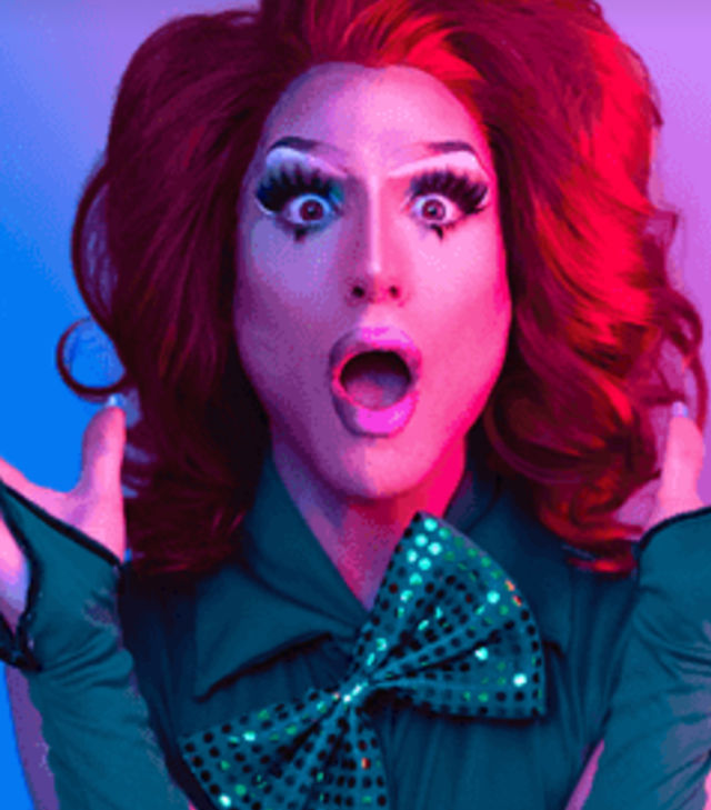 Vibrant portrait of a drag performer with a voluminous red wig, dramatic glam makeup and a surprised open-mouthed expression, wearing a teal outfit with a sequined green bow against neon pink and blue studio lighting.