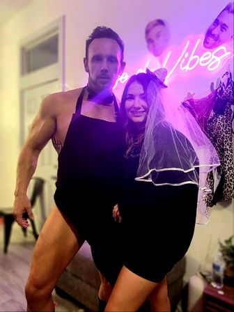 Bachelorette party scene indoors — muscular man in a black apron and bow tie poses with a smiling woman in a short black dress and bridal veil under a pink neon Vibes sign.