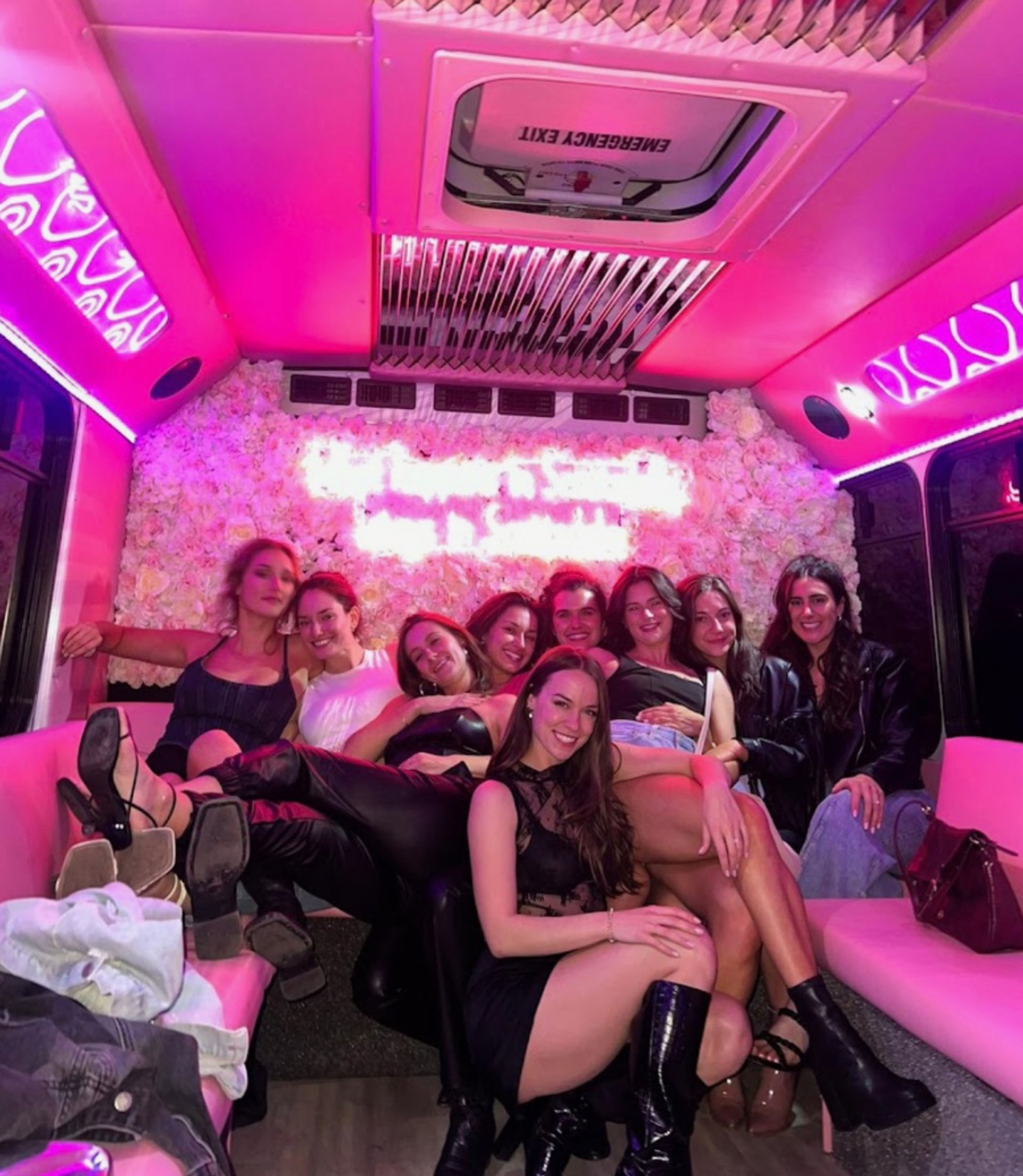 Group of friends lounging in a pink-lit party bus with a flower wall and neon sign, dressed up for a fun night out