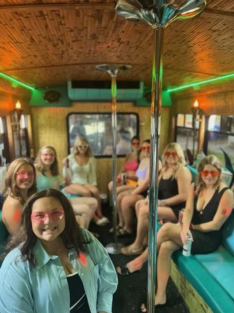 Group of smiling women wearing pink/orange sunglasses seated on turquoise benches inside a party bus, chrome dance poles, bamboo-style ceiling and green neon lighting.