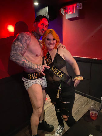 Shirtless tattooed male performer in briefs posing with smiling woman with orange hair wearing a 'Hot Mess' sash in a red-lit nightclub bar interior under an EXIT sign, drinks on a nearby table, party vibe.