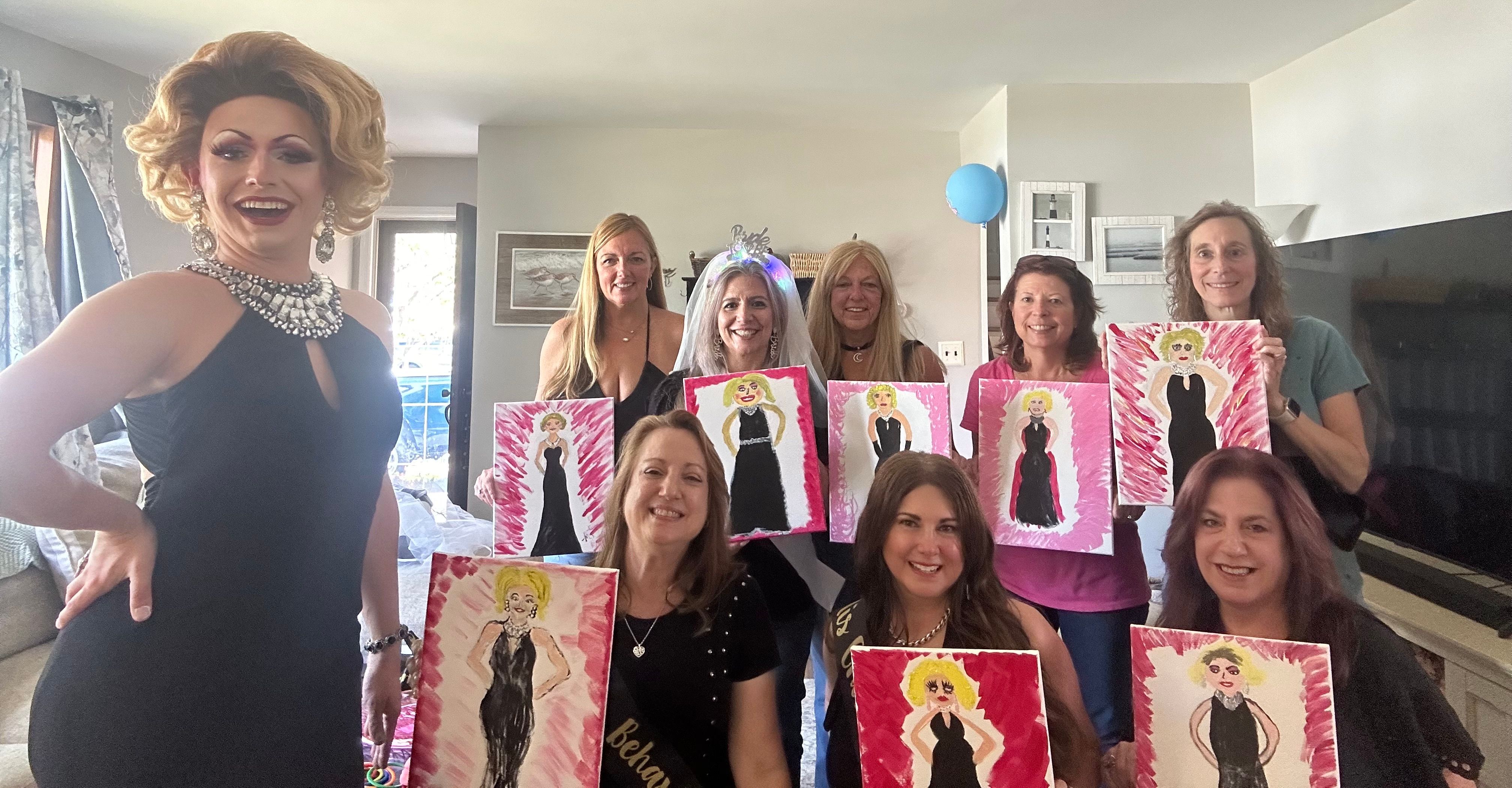 Drag performer in a black gown posing with a group of smiling women in a living room at a paint-and-sip bachelorette party, each holding a canvas of a blonde figure in a black dress; bride-to-be wearing a light-up veil.