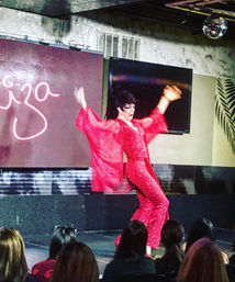 Energetic drag performer in a sparkling red jumpsuit with flowing sleeves striking a pose on a cabaret stage under a disco ball while a seated audience watches.