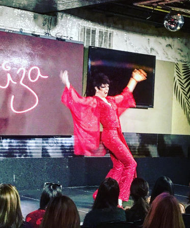 Energetic drag performer in a sparkling red jumpsuit with flowing sleeves striking a pose on a cabaret stage under a disco ball while a seated audience watches.