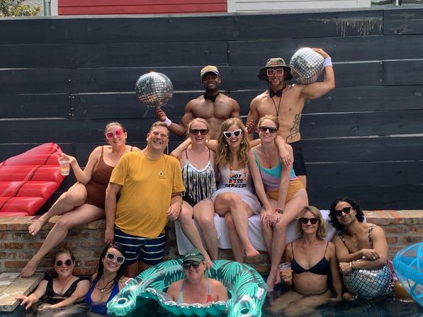 Group of friends at a backyard pool party, adults in swimsuits posing on a brick ledge and in the water with disco ball props, an inflatable float, sunglasses, and summer vibes.