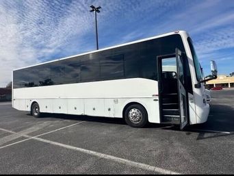 Sleek white charter coach bus with tinted windows and open front door parked in a large asphalt parking lot under a blue sky with wispy clouds.