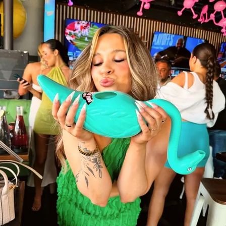 Woman in green dress playfully kissing a turquoise flamingo-shaped novelty drink vessel at a lively sports bar with TV screens and patrons in the background