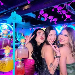 Three friends in glam dresses posing and smiling at a neon-lit, tropical-themed nightclub bar with colorful cocktail dispensers and vibrant pink-blue lighting