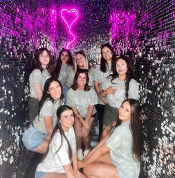 Group of women in matching light gray bachelorette shirts posing and smiling inside a narrow sequin-walled photo booth under a neon pink heart sign — fun party photo