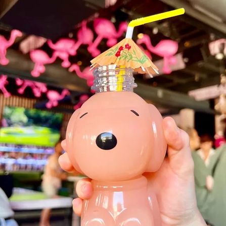 Close-up of a hand holding a pink dog-shaped novelty drink bottle topped with a yellow straw and paper umbrella in a tropical-themed bar with hanging pink flamingo décor
