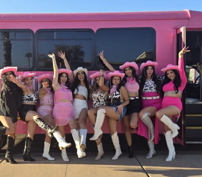 Ten women in pink cowgirl hats, fringed and cow-print outfits and white boots posing cheerfully with one leg raised in front of a bright pink party bus on a sunny day.