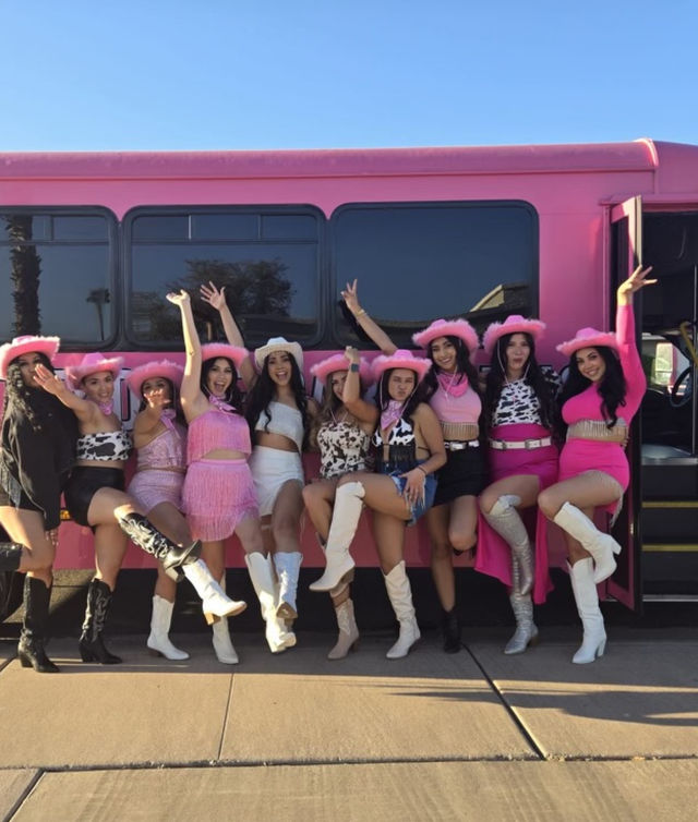 Ten women in pink cowgirl hats, fringed and cow-print outfits and white boots posing cheerfully with one leg raised in front of a bright pink party bus on a sunny day.
