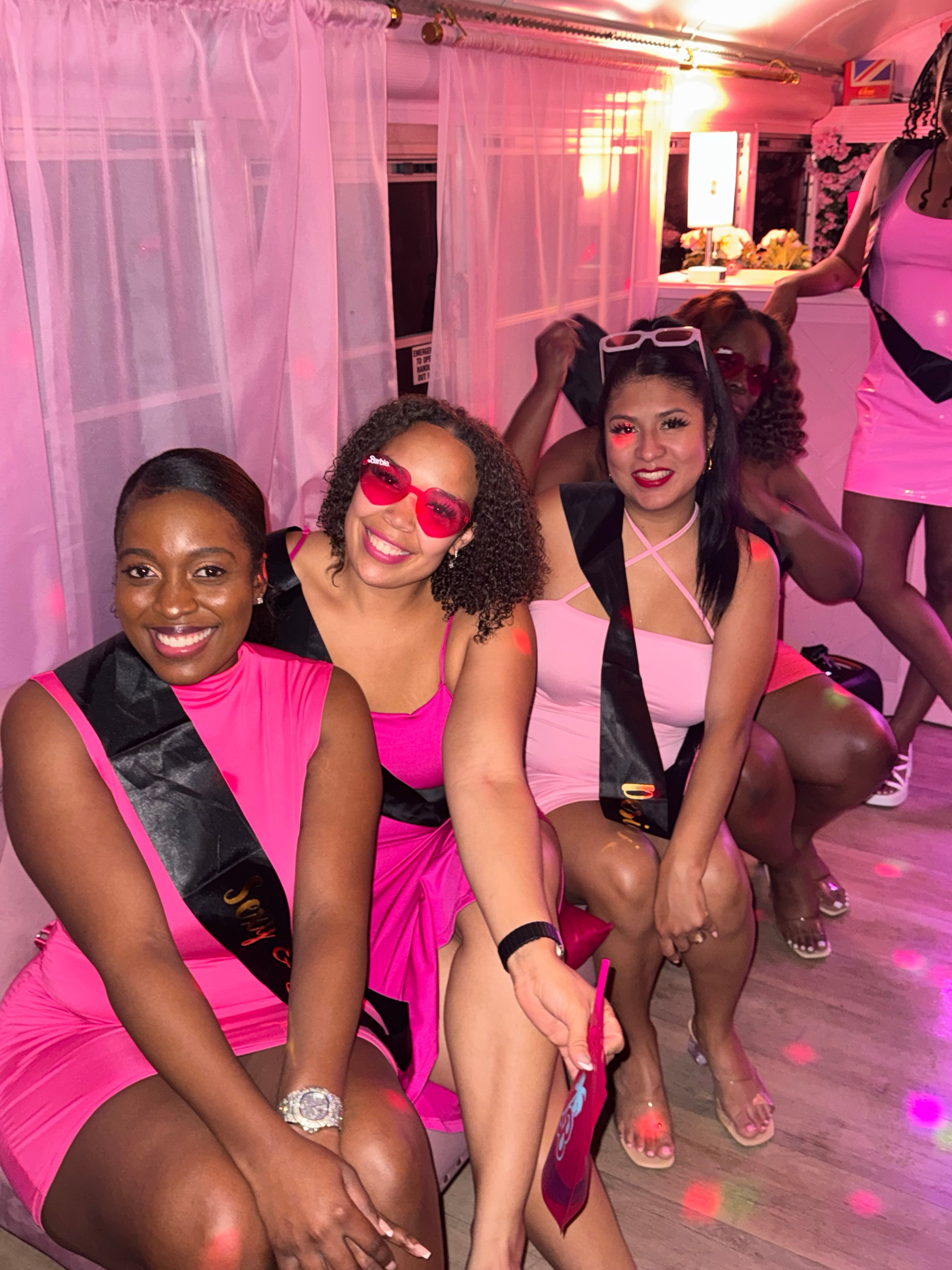 Four friends in pink dresses and black sashes smiling inside a pink-lit party interior, wearing fun sunglasses for a bachelorette/girls' night celebration.