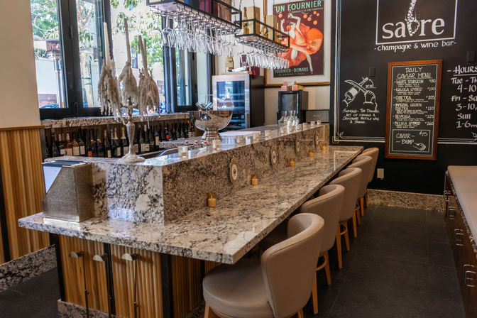 Sleek marble champagne and wine bar interior with rows of hanging wine glasses, plush bar stools, a silver ice bucket on the stone counter and a chalkboard caviar menu by street-facing windows.