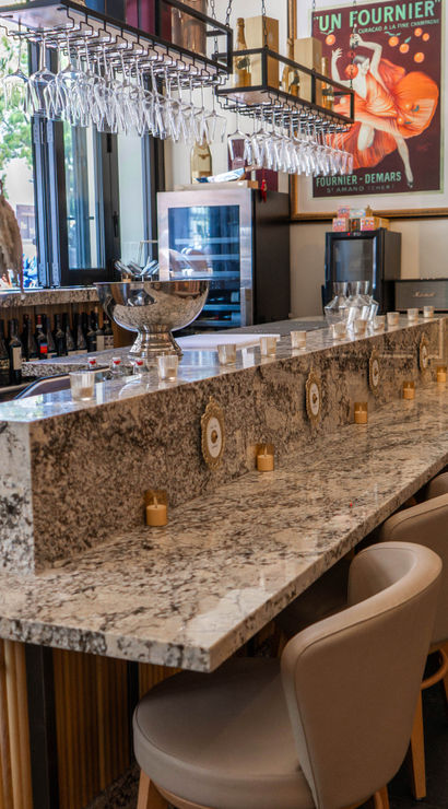 Sleek marble champagne and wine bar interior with rows of hanging wine glasses, plush bar stools, a silver ice bucket on the stone counter and a chalkboard caviar menu by street-facing windows.