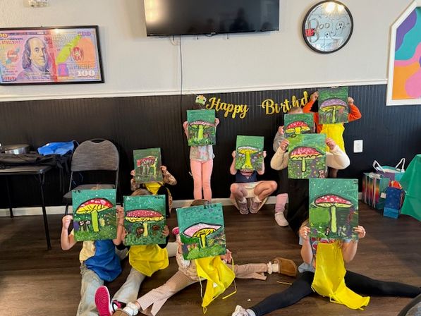 Group of children at an indoor kids' painting birthday party proudly holding colorful mushroom canvas paintings up to hide their faces in a decorated party room