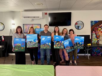 Six people in an indoor art studio holding colorful canvases of blue-sky mountain scenes with whimsical flowers after a group painting class.