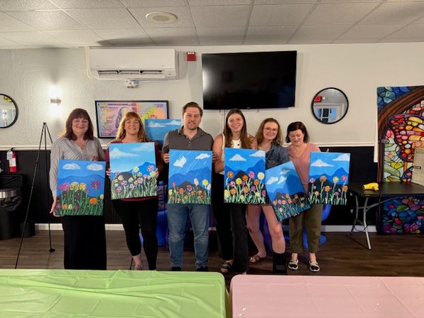 Six people in an indoor art studio holding colorful canvases of blue-sky mountain scenes with whimsical flowers after a group painting class.