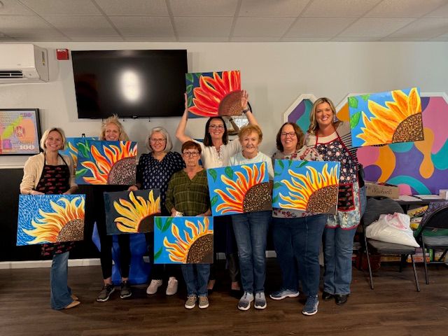 Nine women smiling in an indoor art studio during a paint night, each holding a colorful sunflower canvas painting