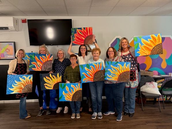 Nine women smiling in an indoor art studio during a paint night, each holding a colorful sunflower canvas painting
