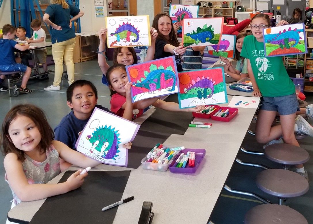 Elementary school children in a classroom art activity proudly holding colorful marker-made dinosaur drawings at a long table with trays of markers and art supplies.