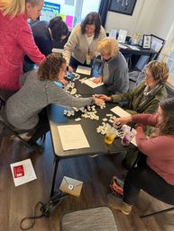 Adults gathered around a folding table in a bright community room sorting dozens of small white game tiles and papers during a collaborative tabletop word-game workshop, with iced drinks and bags nearby.