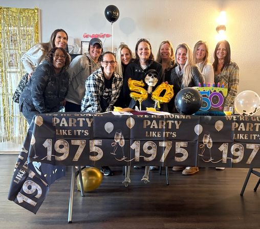Group of smiling women posing behind a decorated table at an indoor 50th birthday party with a 'Party like it's 1975' tablecover, gold '50' balloons, black and gold balloons, colorful cake sign and shimmering backdrop