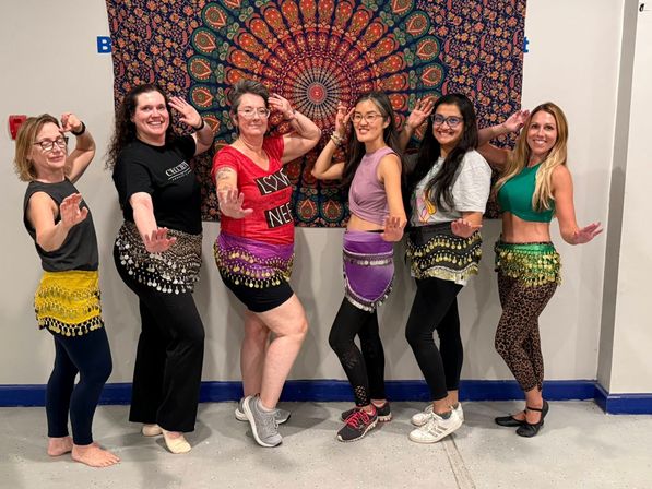 Group of seven women in a belly-dance class posing in a studio, wearing colorful coin hip scarves and athletic wear in front of a vibrant mandala tapestry.