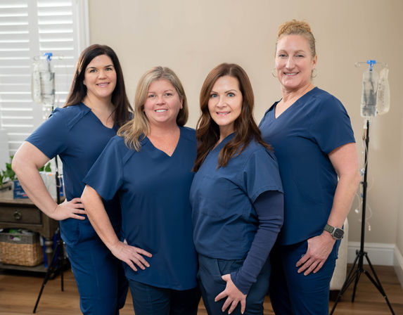Four smiling female healthcare professionals in navy scrubs standing in a bright clinic room with IV drip stands — friendly medical team for IV therapy and wellness.