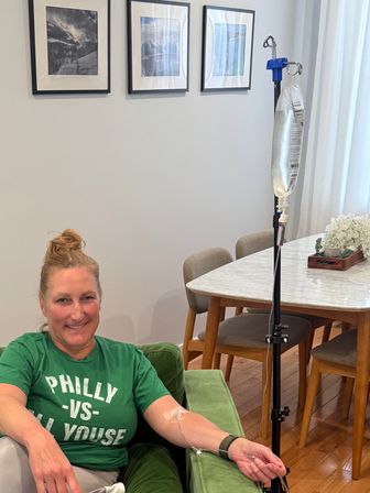 Smiling woman in a green 'PHILLY VS' shirt seated on a green couch receiving an IV infusion at home, IV pole and bag beside a marble-topped dining table with framed photos on the wall.