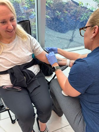 Smiling woman seated by a window receiving a blood draw from gloved medical staff in a bright community health clinic, with an arm tattoo visible.