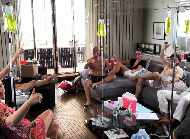 Group of adults relaxing in a boat or yacht lounge with hanging IV infusion bags on poles, smiling, giving thumbs up, casual summer clothes, wooden floor and open balcony doors