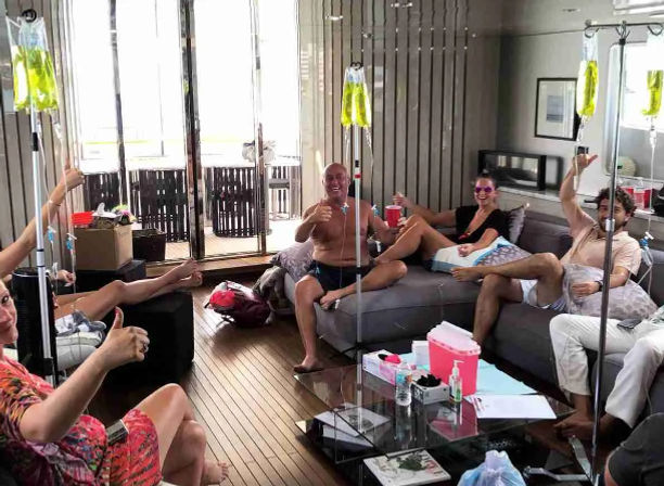 Group of adults relaxing in a boat or yacht lounge with hanging IV infusion bags on poles, smiling, giving thumbs up, casual summer clothes, wooden floor and open balcony doors