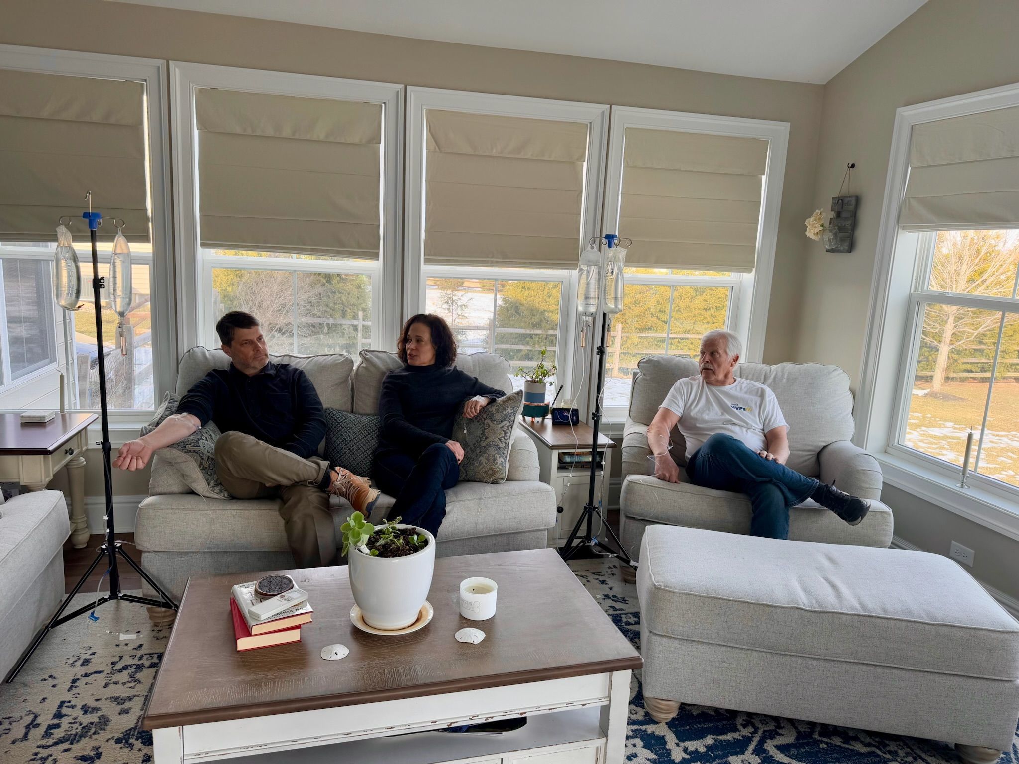 Sunlit suburban living room with three adults seated on a light-gray sofa and armchair, two IV drip stands by large windows, wooden coffee table with potted plant and books, neutral decor and patterned rug.