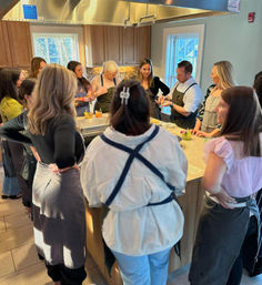 Small-group cooking class gathered around a kitchen island, chef demonstrating a technique while participants in aprons watch and chat in a bright home-style kitchen.