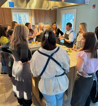 Small-group cooking class gathered around a kitchen island, chef demonstrating a technique while participants in aprons watch and chat in a bright home-style kitchen.